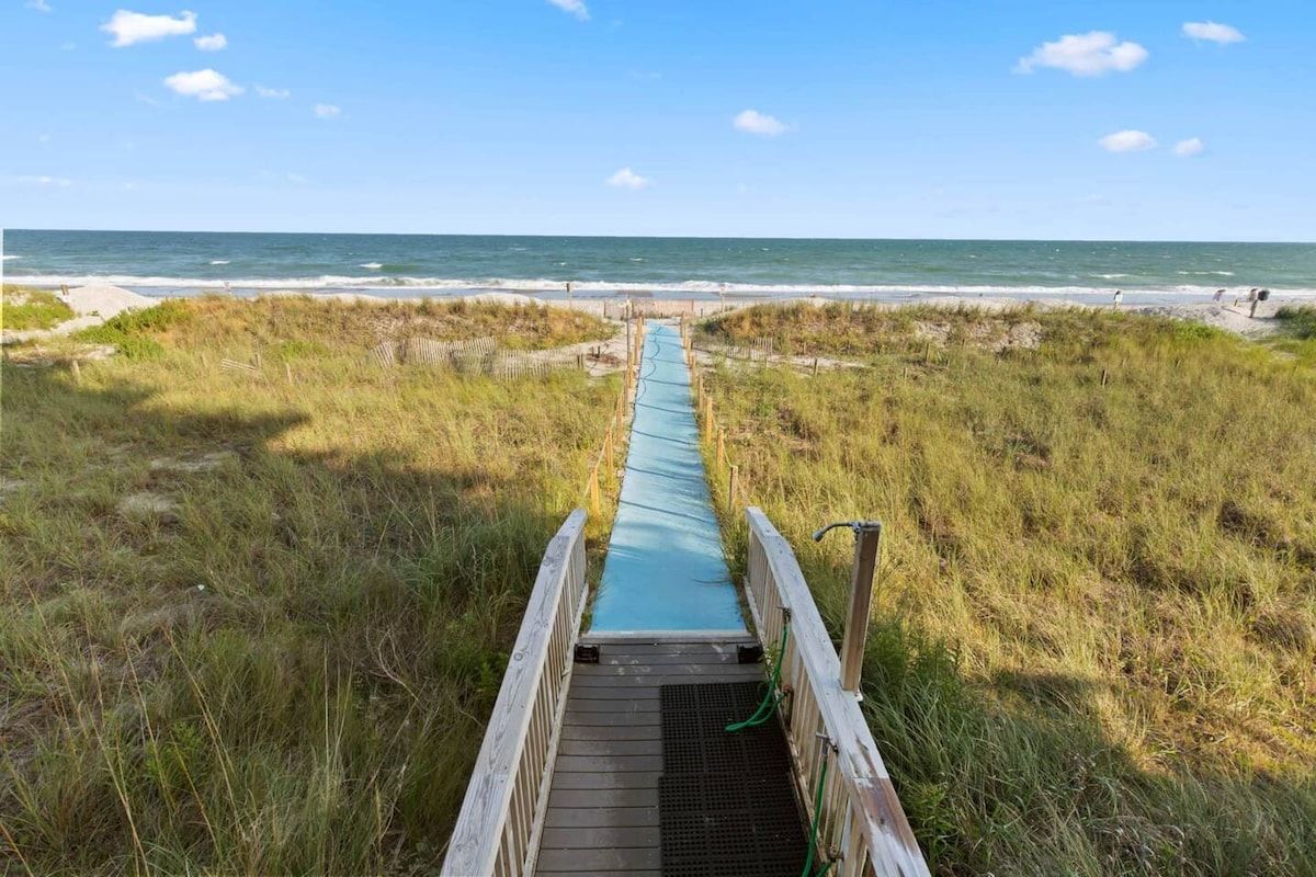 Ein Holzsteg führt zu einem Strand unter blauem Himmel.