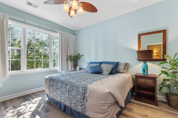 Une chambre à coucher avec un grand lit, un ventilateur de plafond, une fenêtre avec des rideaux et une table de chevet avec une lampe.