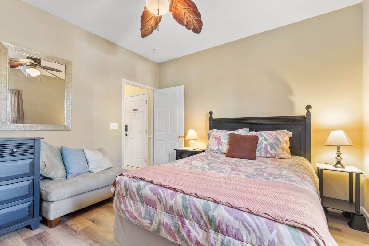 Quarto com cama, cômoda, espelho, porta e mesa de cabeceira com abajur.