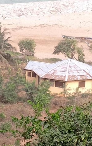 A rural house with a corrugated metal roof, surrounded by vegetation and a sandy beach in the background.