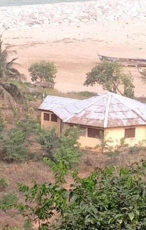 Una casa rural con techo de metal corrugado, rodeada de vegetación y una playa de arena al fondo.