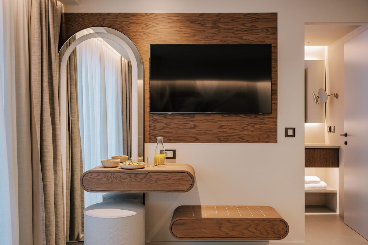A modern bathroom with a wooden vanity, a flat-screen TV mounted on the wall, and a shower area visible through an open door.