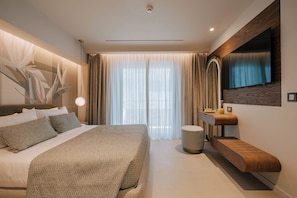 A modern hotel room with a large bed, a built-in bench, a flat-screen TV, and a large window with sheer curtains. - Cento Palme (Rhodes)