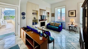 Ein Wohnzimmer mit blauem Sofa, einem Holztisch und einem Fernseher. Es gibt Bücherregale und einen Balkon mit Sitzgelegenheiten.
