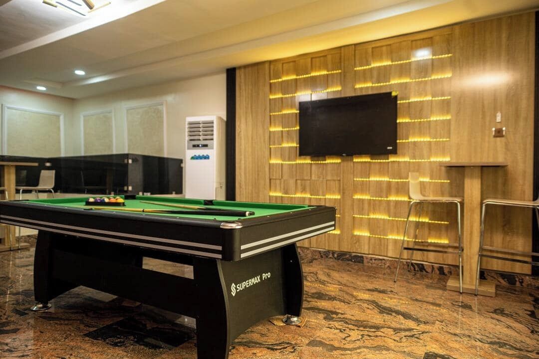 Una mesa de billar con etiqueta Supermax Pro, un televisor de pantalla plana montado en una pared de madera y una zona de estar con sillones.