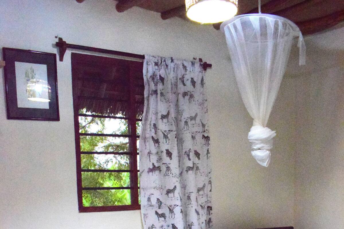 A bedroom with a bed, a chair, a window with curtains, and a hanging mosquito net.