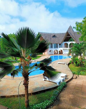 Een resort met een zwembad, palmbomen en een groot huis met rieten dak.