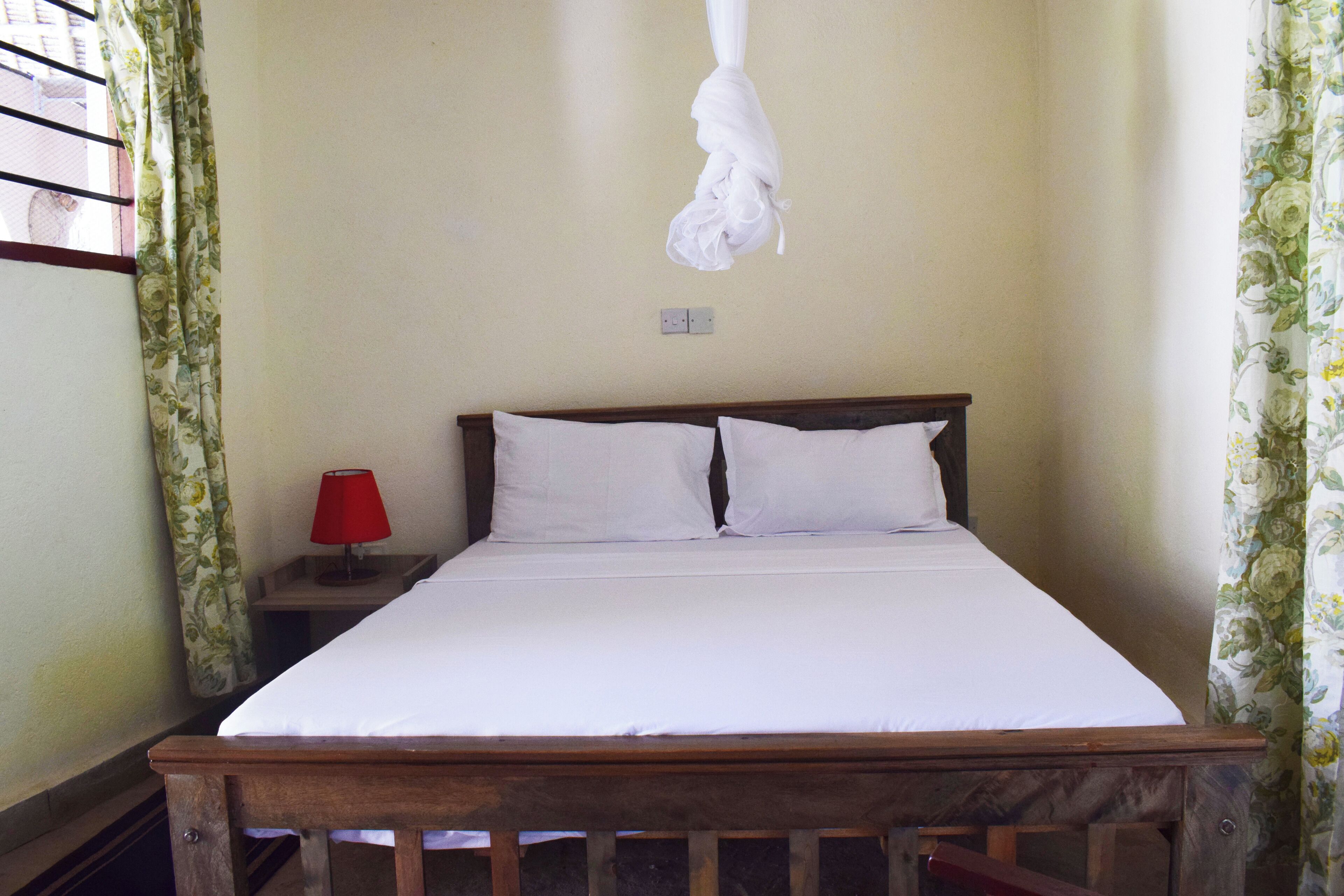 Un dormitorio con cama de madera, ropa de cama blanca, una lámpara roja sobre una mesita de noche y cortinas con estampado floral.