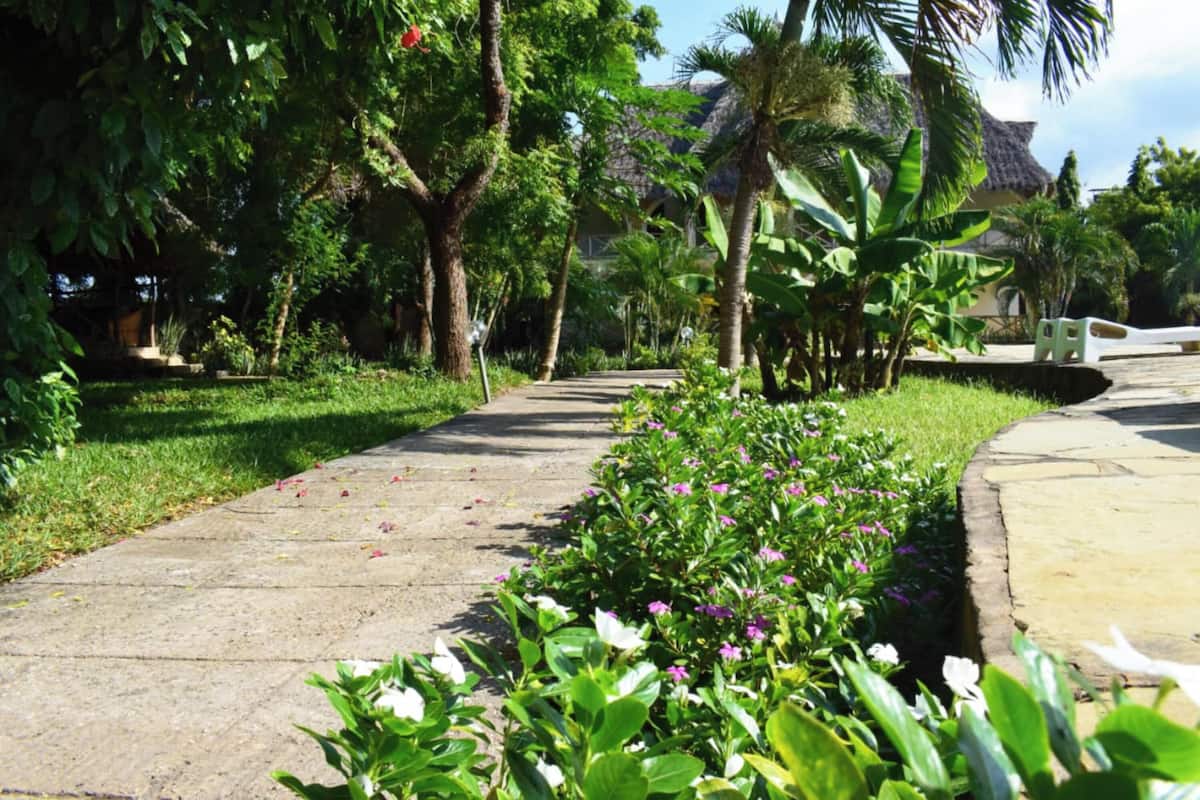 A pathway lined with lush greenery and flowering plants, leading to a building with a thatched roof.