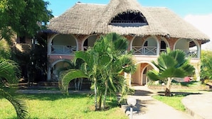 A thatched-roof building with a central courtyard, surrounded by palm trees and a paved walkway. - Raquel's Bliss Hotel Diani (Diani Beach)