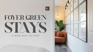 A hotel interior with a wall sign reading "FOYER GREEN STAYS A NEW WAY TO STAY" and a seating area with a plant and framed pictures.