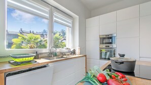 Eine moderne Küche mit Geschirrspüler, Spüle, einem Fenster mit Blick auf Gebäude und Grünflächen sowie einer Arbeitsplatte mit einem Topf und Gemüse.