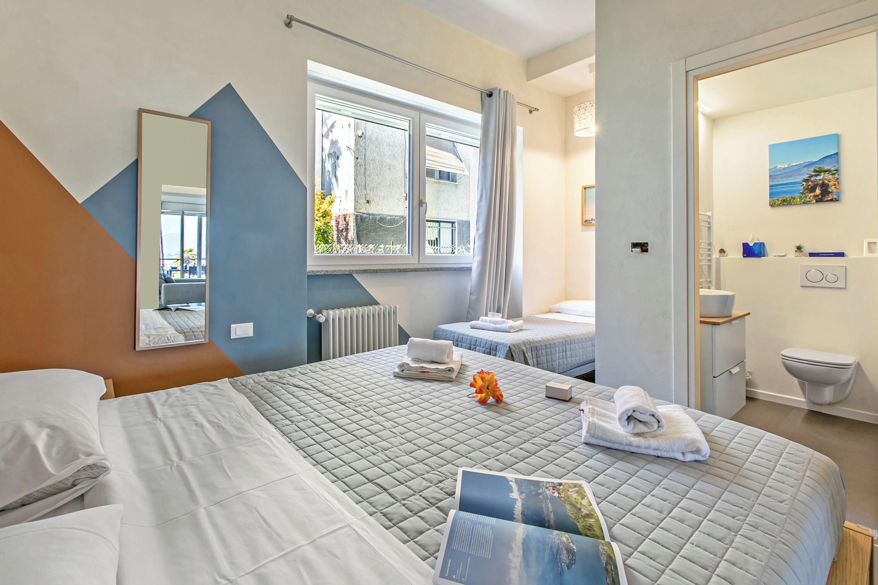 Una camera d'albergo con un letto grande, un letto più piccolo, una finestra con tende, un bagno e un quadro appeso al muro.