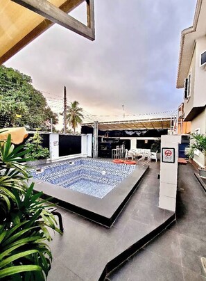 Une piscine extérieure avec un sol carrelé, entourée d’une terrasse en béton et agrémentée de plantes en pot.
