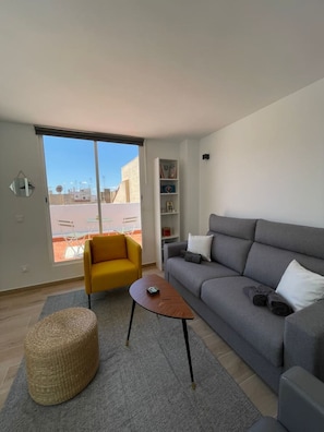 Una sala moderna con un sofá gris, un sillón amarillo, una mesita de centro de madera y un otoman tejido.