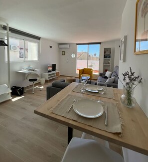 A modern living room with a dining table set for two, a sofa, a yellow armchair, and a view of the cityscape through large windows.