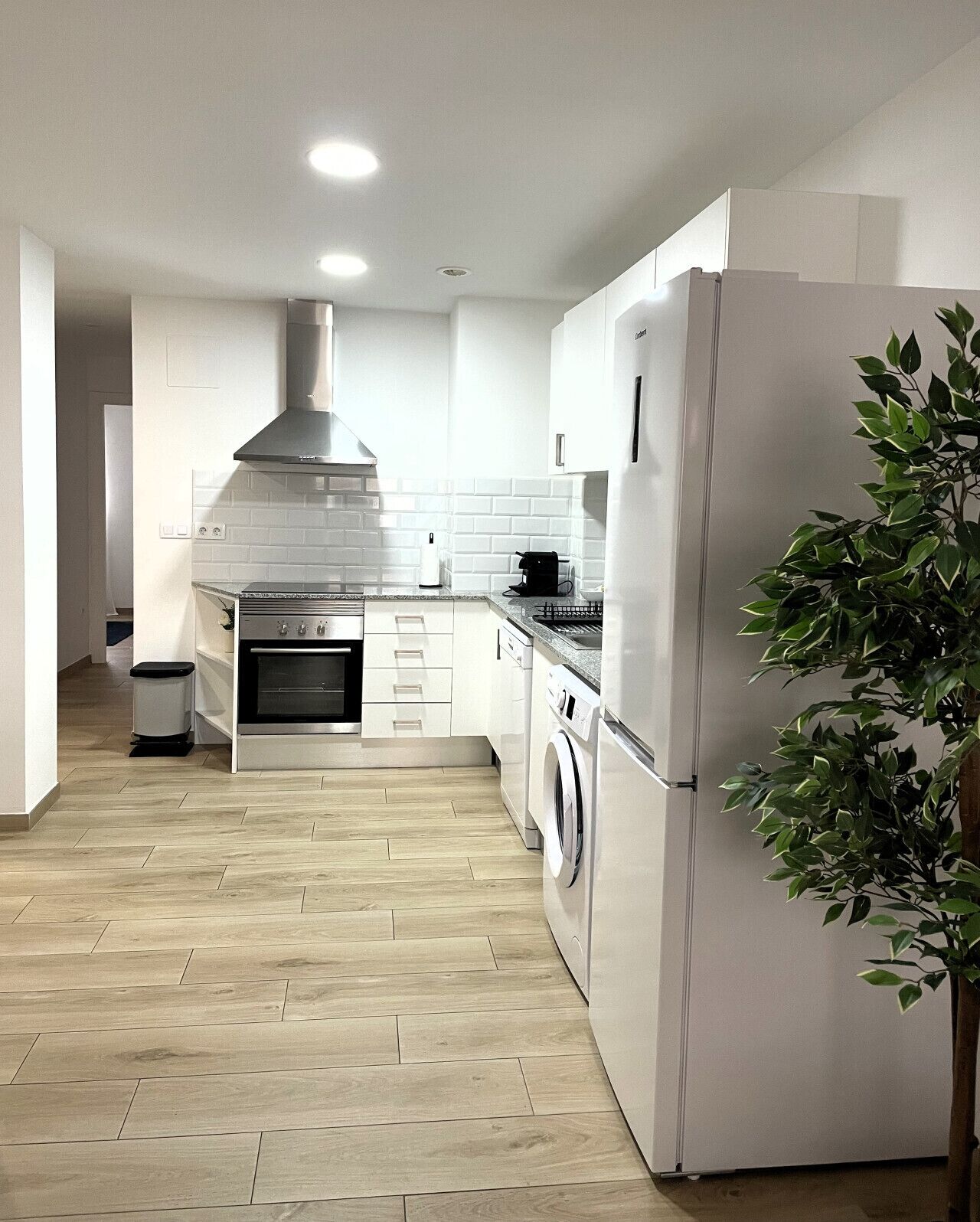 A modern kitchen with white cabinetry, a stainless steel range hood, a built-in oven, a washing machine, and a refrigerator.