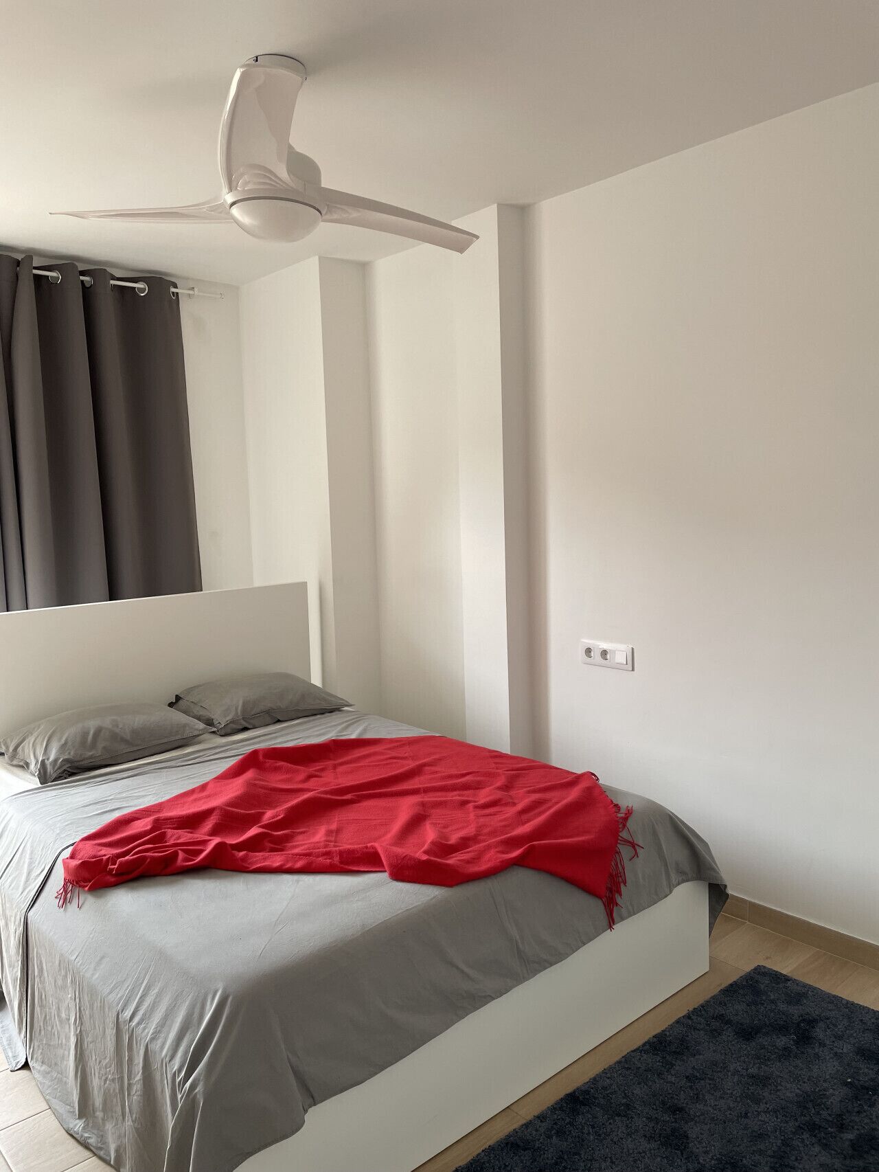 A bedroom with a bed, a ceiling fan, a dark rug, and a light-colored wall.