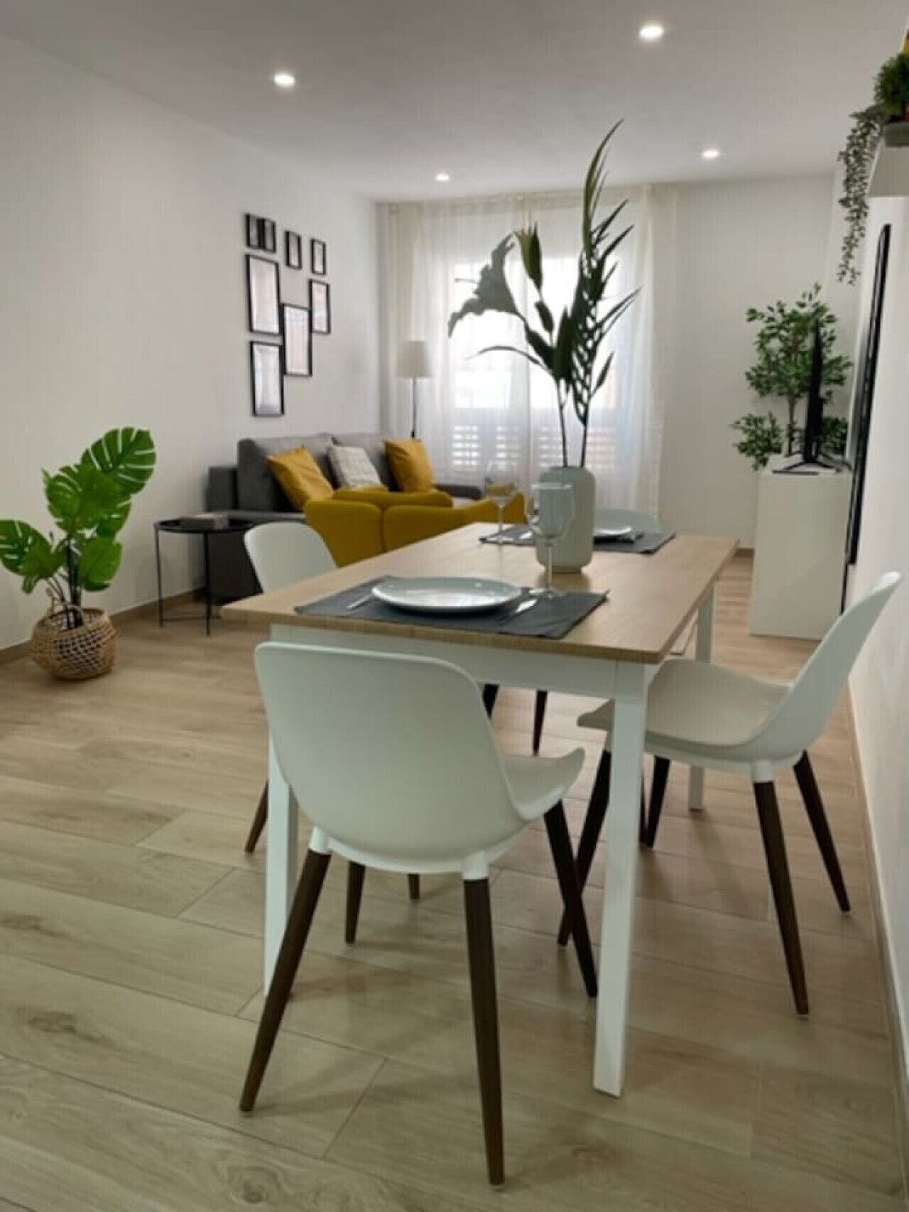 Una sala de estar moderna con una mesa de comedor, sillas blancas, un sofá con cojines amarillos y una planta grande.