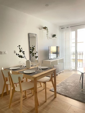 Un comedor moderno con una mesa de madera dispuesta para cuatro, un espejo grande y una pequeña cocina minimalista al fondo.