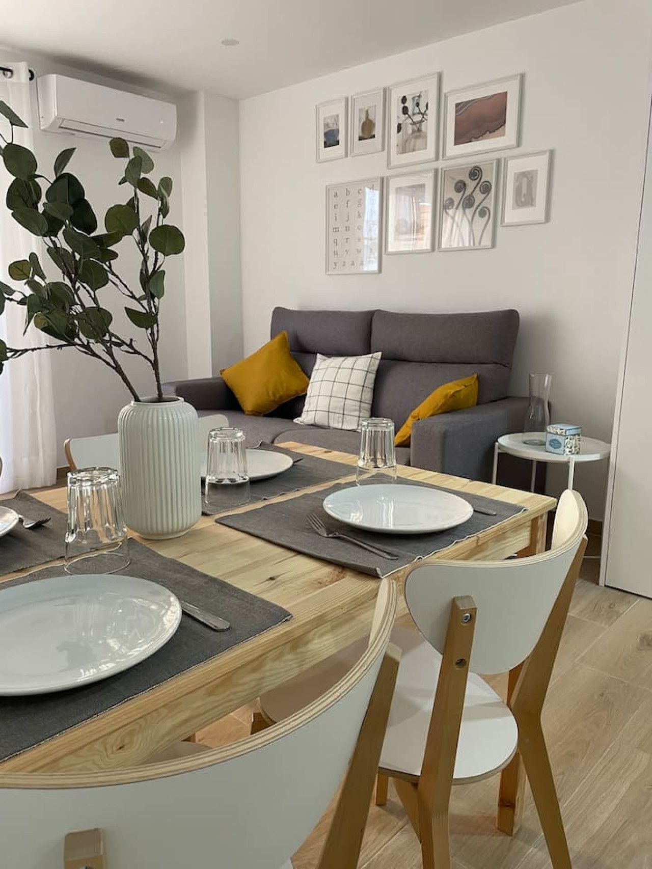 Un comedor con mesa de madera dispuesta para cuatro, un sofá gris con cojines amarillos y un jarrón blanco con hojas verdes.