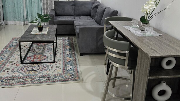 A modern living room with a grey sofa, a coffee table, a patterned rug, and a dining area with a wooden table and chairs. - Comfy Fresh and Clean Spacious 1- bedroom apartment with AC in charming Portmore (Portmore)