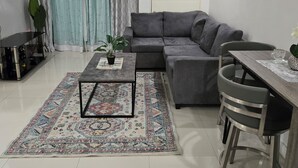 A modern living room with a grey sectional sofa, a coffee table, a dining table with chairs, a TV mounted on the wall, and a large window with patterned curtains. - Comfy Fresh and Clean Spacious 1- bedroom apartment with AC in charming Portmore (Portmore)