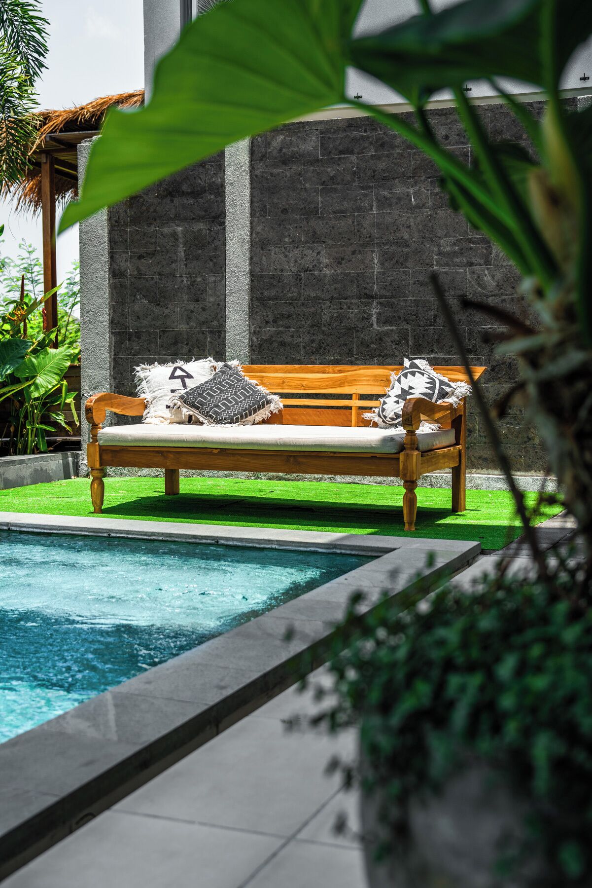 Un banc en bois avec des coussins, placé près d’une piscine, entouré de verdure et bordé d’un mur de pierre.