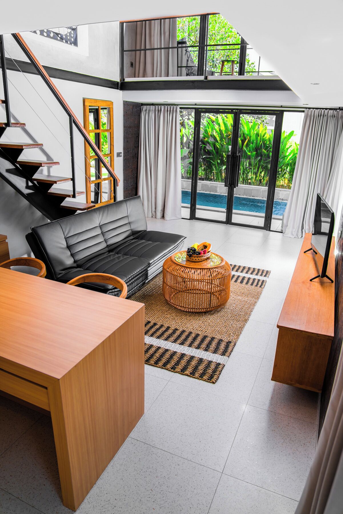 Uma sala de estar moderna com um sofá de couro preto, uma mesa de centro de madeira e uma TV. Há uma escada que leva a um andar superior e uma vista para uma piscina externa.