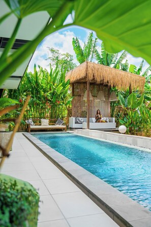 Un espace piscine agrémenté d’un abri au toit de chaume, de chaises longues et d’une végétation tropicale luxuriante.