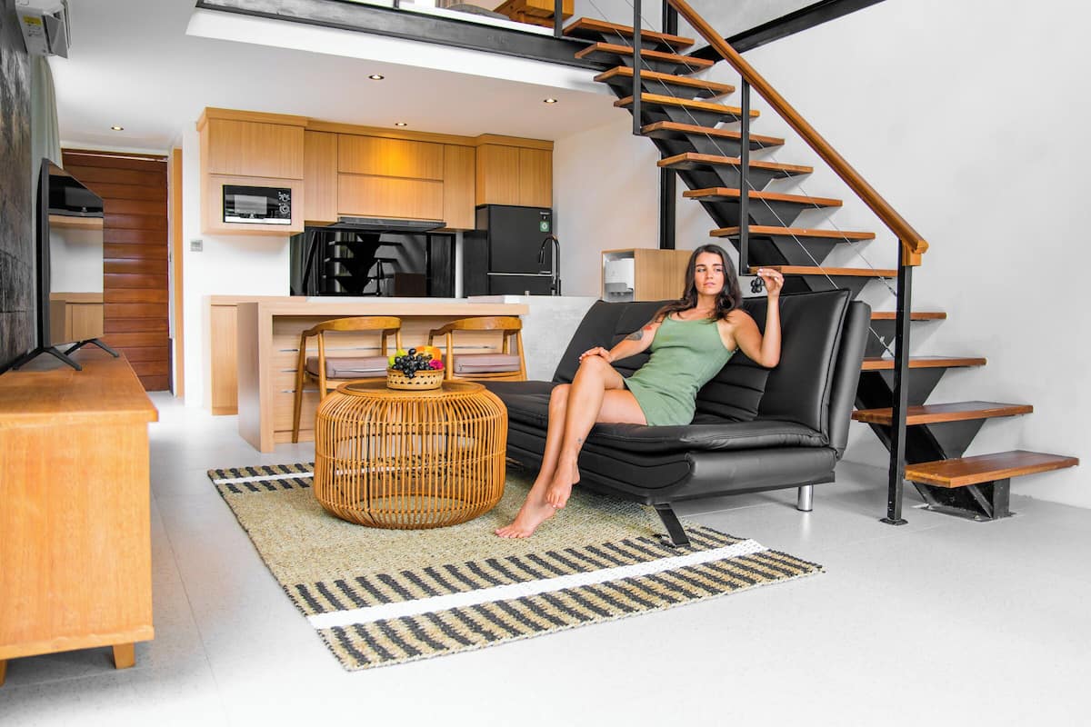 A modern living room with a black sofa, wooden staircase, and a kitchen area in the background.