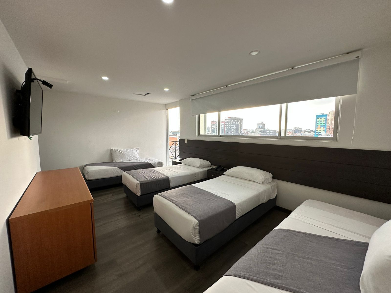 A hotel room with two single beds, a double bed, a wooden side table, a flat-screen TV mounted on the wall, and a large window with a city view.