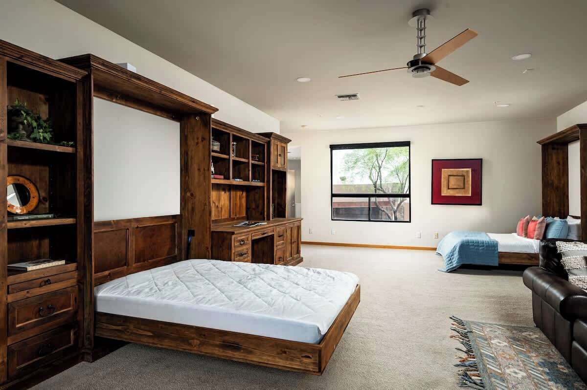 Un dormitorio con estructura de cama de madera, un escritorio con estanterías, un ventilador de techo, una ventana con vistas y una cama con manta azul.
