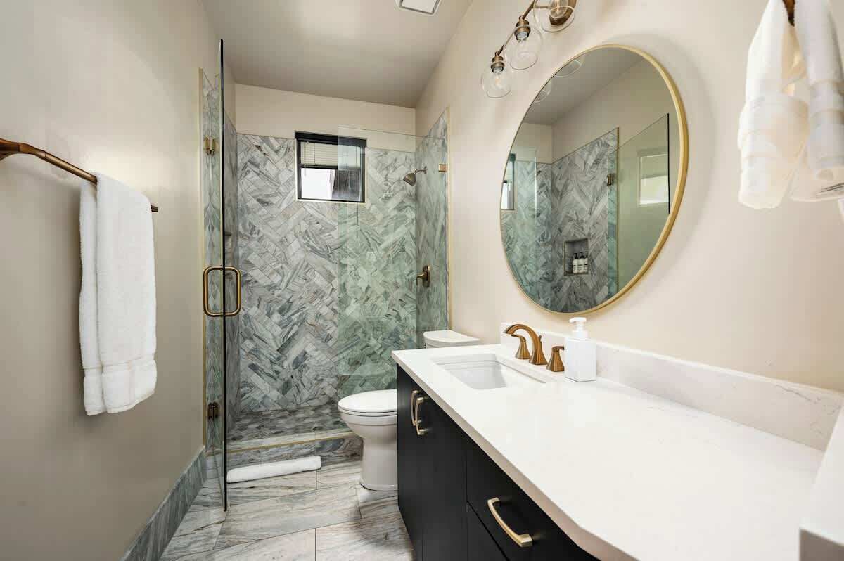 Un baño moderno con cabina de ducha de vidrio, lavamanos blanco con detalles dorados y un espejo grande.