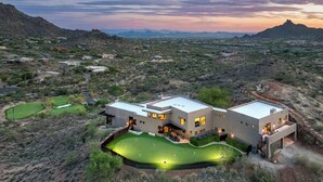 A modern two-story house with a flat roof, surrounded by a desert landscape, with a well-maintained lawn and a golf course nearby.