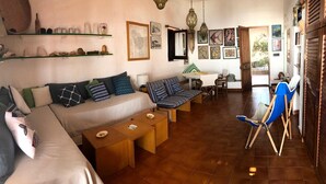 Ein gemütliches Wohnzimmer mit einer Couch, Sesseln und einem Esstisch. An den Wänden befinden sich Regale mit Dekoration und Kunstwerken.