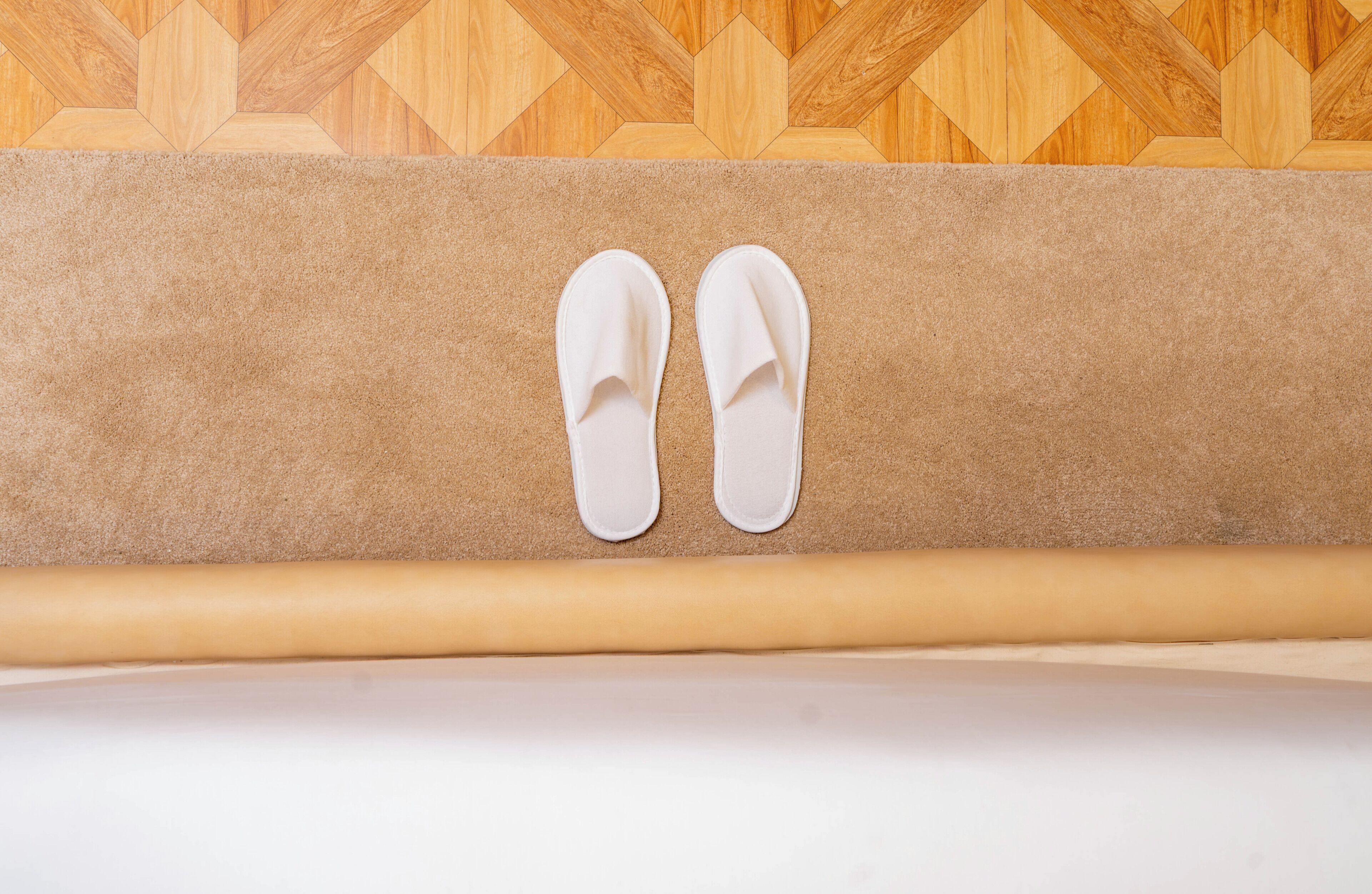 A pair of white slippers on a beige carpet, a wooden patterned wall, and a white floor.