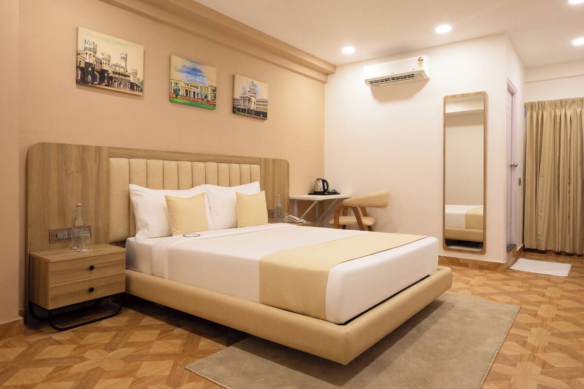 A modern hotel room with a large bed, a bedside table with a lamp, a small desk, and a mirror.