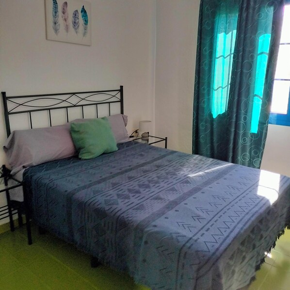 Un lit avec une couvre-lit à motifs, un oreiller vert et un oreiller rose. Au-dessus du lit, on aperçoit une œuvre d’art accrochée au mur, et à droite, une fenêtre avec des rideaux.