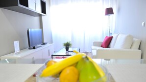 A modern living room with a glass coffee table in the foreground, a white sofa with a red pillow, a flat-screen TV on a white cabinet, and a potted plant on a glass coffee table. - Goya - 1 Bedroom Apartment (Madrid)