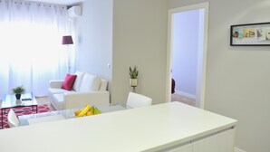 A modern living room with a white sofa, a glass coffee table with fruits, and a small plant. - Goya - 1 Bedroom Apartment (Madrid)