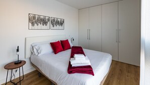 Ein modernes Schlafzimmer mit einem großen Bett, weißer Bettwäsche, roten Kissen und einer roten Tagesdecke. Über dem Bett befinden sich zwei Wandbilder und ein kleiner Holznachttisch mit einer Lampe.
