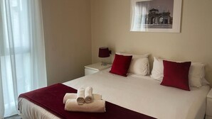 Une chambre d’hôtel avec un lit, une table de chevet, une lampe, un tableau encadré au mur et une fenêtre avec des rideaux légers.