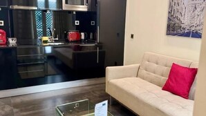 Une cuisine moderne avec des meubles de couleur sombre et des appareils électroménagers en acier inoxydable, une table basse en verre et un canapé beige agrémenté d’un coussin rouge.