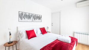 Una cama bien hecha con ropa de cama blanca y roja, una manta roja y una toalla blanca al lado. Hay tres obras de arte colgadas en la pared sobre la cama, una mesita redonda con una lámpara junto a la cama y un radiador blanco en la pared.