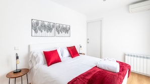 A neatly made bed with white and red bedding, a red throw blanket, and a white towel on the side. There are three wall-mounted artworks above the bed, a small round table with a lamp beside the bed, and a white radiator on the wall.