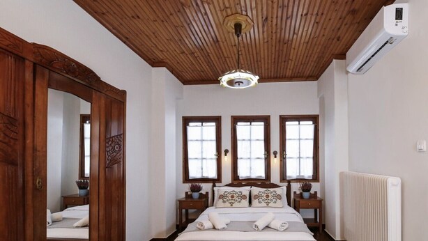 Un dormitorio con techo de madera, un espejo grande, tres ventanas, una cama, mesitas de noche y un radiador.
