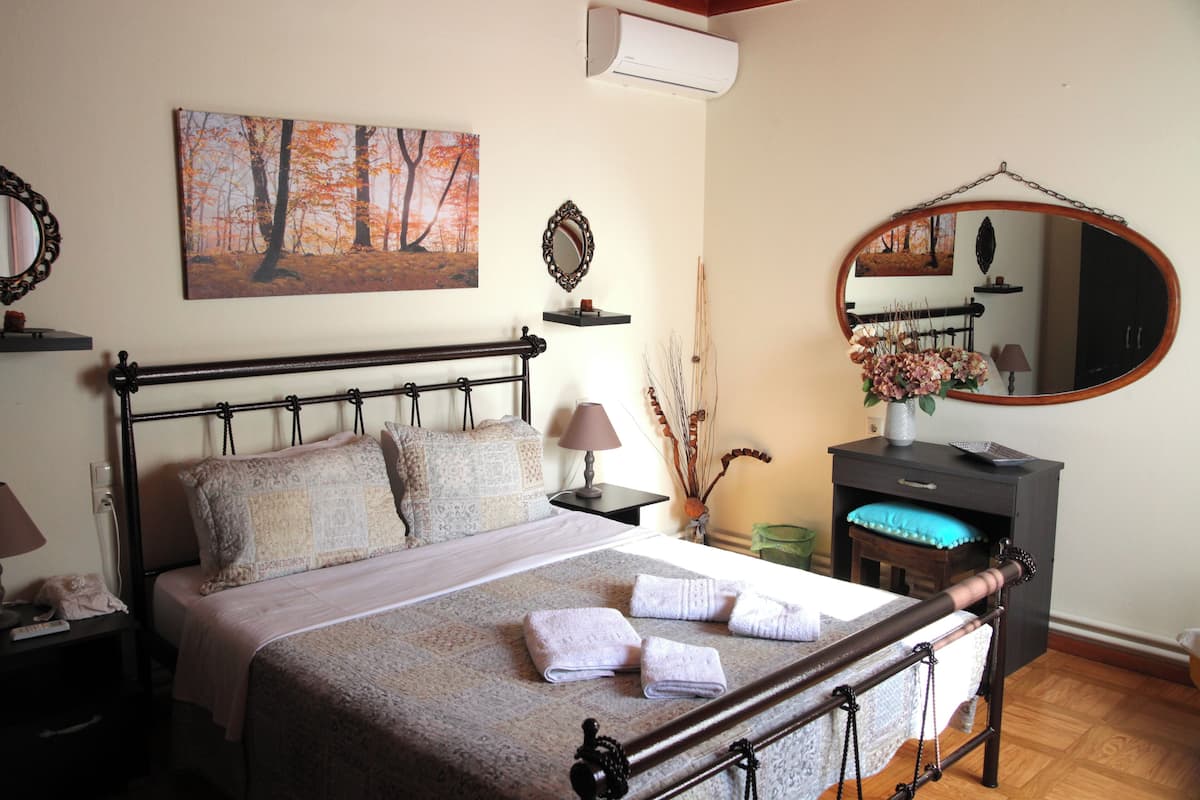 A neatly made bed with pillows and towels, a bedside table with a lamp, a mirror, and a painting of trees on the wall.