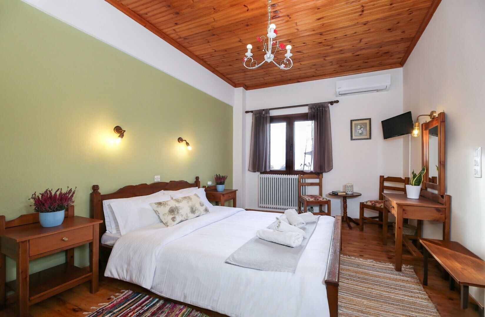 A bedroom with a wooden ceiling, a bed with white linens, a bedside table with a plant, a chair, a desk with a mirror, and a television on the wall.