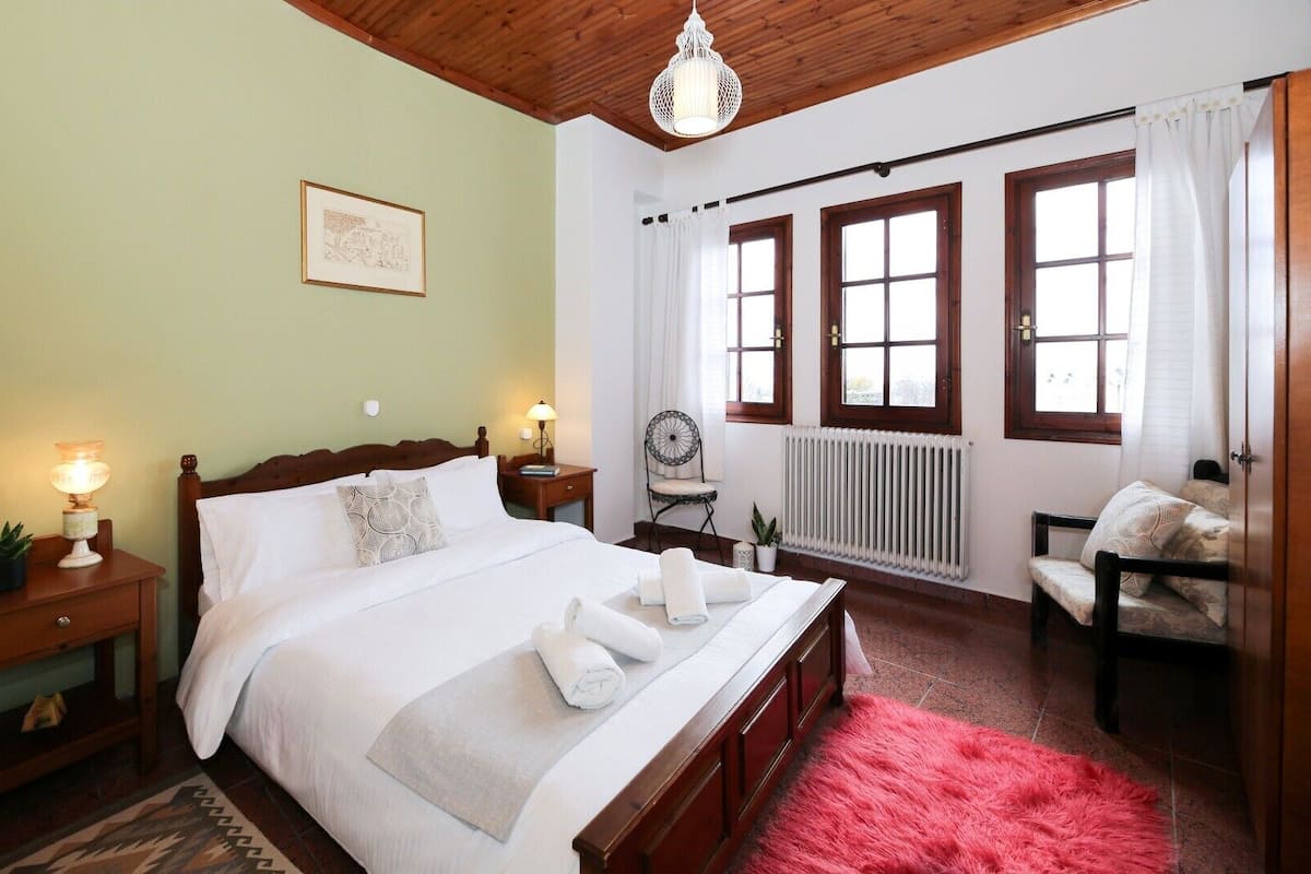 A neatly arranged bedroom with a wooden bed, bedside lamps, a radiator, a chair, and a window with curtains.